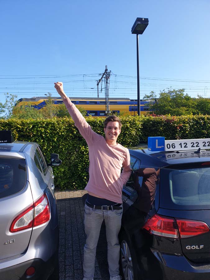 Geslaagde leerling met duimen omhoog naast de lesauto van Rijschool Amsterdam
